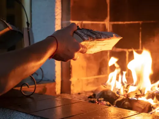 En hand lägger ved i en sprakande brasa. Det syns tecken på soteld i eldstaden.