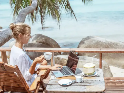 Kvinna med kaffe och laptop på en strandterrass, drömmen om ett digitalt nomad jobb.