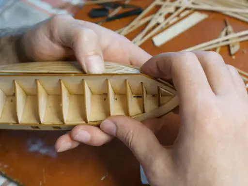 Hände bauen einen hölzernen Schiffsrumpf. Die Rippen des Kaders sind sichtbar, während die äußere Hülle geformt wird.