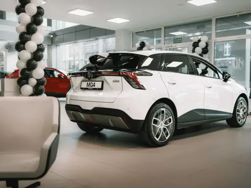 Białe auto MG 4 Electric w salonie, ozdobionym balonami. Obok czerwony samochód i wygodne fotele.