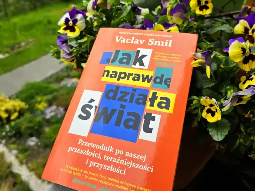Książka Vaclava Smila "Jak naprawdę działa świat" wśród kwiatów. To przewodnik po przeszłości, teraźniejszości i przyszłości, który pomaga zrozumieć, co to odwyk od błędnych przekonań.