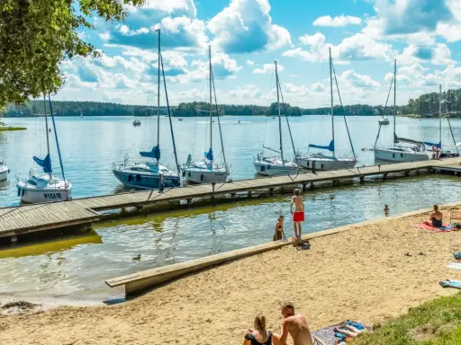 Słoneczny dzień nad jeziorami mazurskimi. Drewniane molo z żaglówkami, piaszczysta plaża z wypoczywającymi ludźmi i błękitne niebo z chmurami.