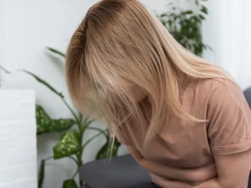 Kobieta z blond włosami siedzi, trzymając się za brzuch. Wygląda na to, że cierpi z powodu zatrucia pokarmowego.