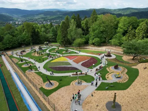 Widok z góry na park z kolorowymi rabatami kwiatowymi i rzeźbami. Widać ścieżki, ławki i ludzi spacerujących po terenie. W tle góry i las.