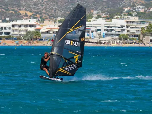Ein Mann beim Windsurfen in Griechenland, mit Blick auf eine belebte Küste und Berge im Hintergrund.
