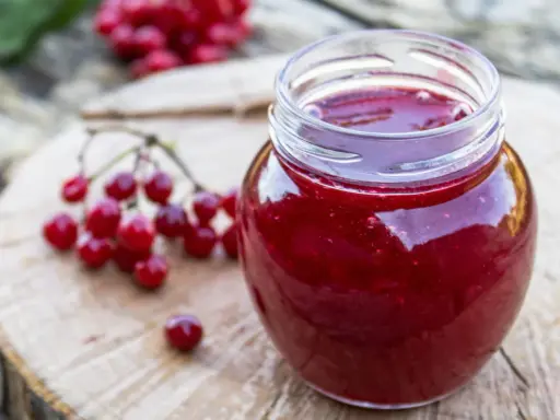 Un pot de gelee de groseille, aux reflets rubis, est posé sur une planche de bois. Des grappes de groseilles rouges sont disposées à côté.
