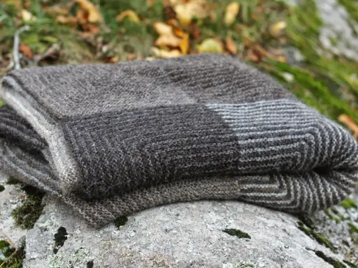 Vikta plädar i ull med mönster som påminner om dominostickning, ligger på en mossig sten utomhus.