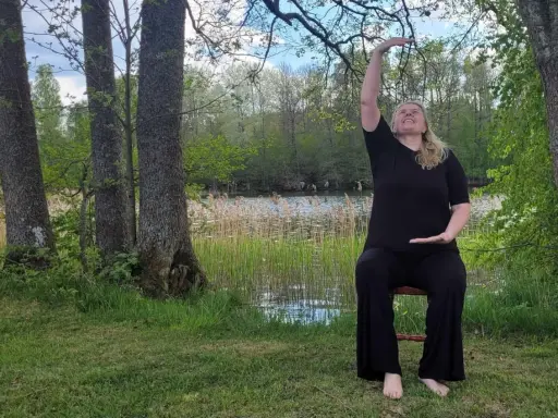 Kvinna i svart klädsel utför yoga på en stol vid en sjö. Naturen är grönskande med träd och vass.