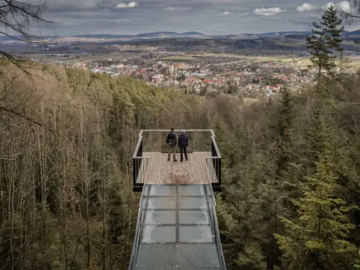 Dwie osoby stoją na szklanym tarasie widokowym, podziwiając panoramę Starego Sącza i okolic.