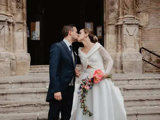 Novios besándose frente a una iglesia, ella lleva un ramo de flores. Un momento especial tras las lecturas de boda religiosa.