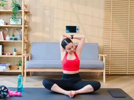 Frau dehnt sich im Schneidersitz, ein wichtiger Teil ihres Mobility Trainings. Trainingsmatte, Wasserflasche und Hanteln liegen bereit.