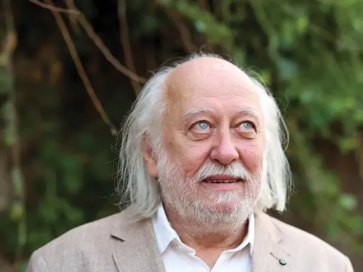 Ritratto di Laszlo Krasznahorkai, scrittore ungherese, con capelli bianchi e barba, sguardo pensieroso rivolto verso l'alto.