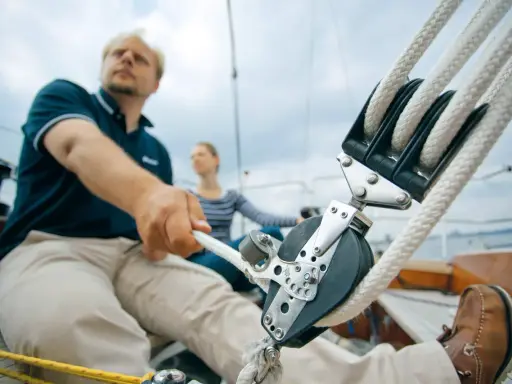 Mann zieht mit kräftiger Hand das Tauwerk am Segel. Ein Mann und eine Frau auf einem Segelboot, die das Tauwerk am Segel bedienen.