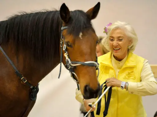 Linda Tellington mit einem braunen Pferd, sie trägt eine gelbe Weste und eine Blume im Haar.
