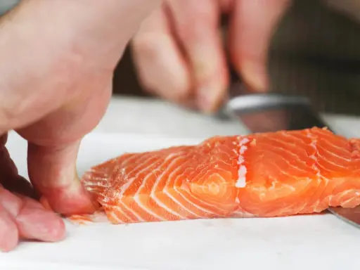 Hände halten ein Lachsfilet, während ein Messer die Haut vom Lachs entfernt.