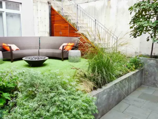 Un coin béton transformé en jardin : canapé, feu de camp, plantes luxuriantes et un chien curieux.