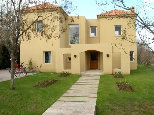 Una encantadora casa color arena con techo de tejas rojas, un camino de losas y dos bicicletas aparcadas en el jardín.