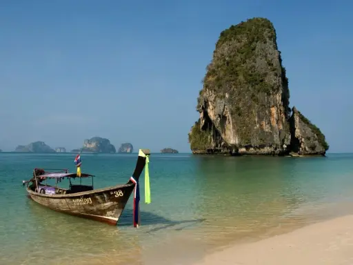 En traditionell longtailbåt guppar i det turkosa vattnet vid en strand i Thailand. I bakgrunden reser sig en grönskande klippö.