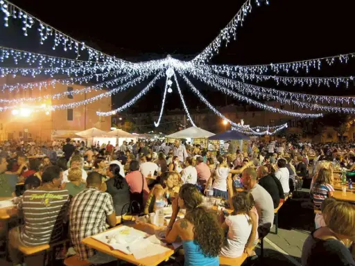 Festa notturna con migliaia di persone sedute ai tavoli, illuminate da festose luminarie. Si celebra la sagra te lu purpu, un evento conviviale.