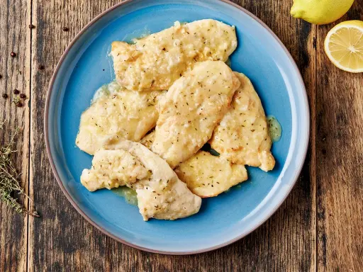 Piatto di pollo al limone su un piatto blu, con limone fresco e rosmarino sullo sfondo.