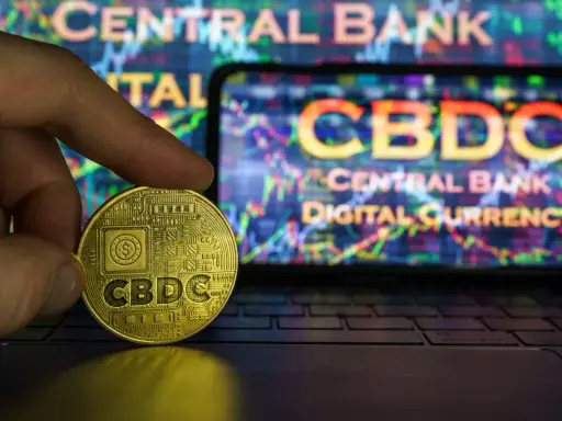Złota moneta z napisem CBDC w dłoni, na tle ekranu z wykresem i napisem "Central Bank Digital Currency".