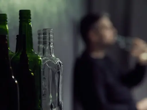 Puste butelki na pierwszym planie, w tle niewyraźna postać pijąca alkohol. Gdzie jest bezpieczna ilość alkoholu?