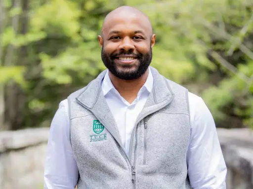 A smiling man with a beard wears a grey vest over a white shirt. The vest has a Tuck at Dartmouth logo, suggesting a curated professional image.