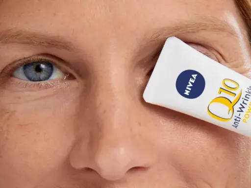 Un tube de crème pour les yeux Nivea Q10 Anti-Wrinkle Power est tenu devant l'œil d'une femme, masquant les rides pattes d'oie.