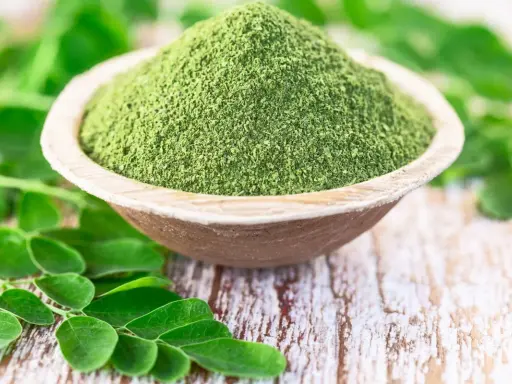 Moringa w proszku w drewnianej misce, otoczona świeżymi liśćmi. Zanim zaczniesz stosować, sprawdź moringa przeciwwskazania.