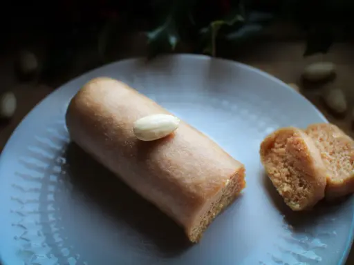 Un bâtonnet de pâte d'amande, garni d'une amande entière, repose sur une assiette blanche. À côté, deux tranches de ce délice, parfait pour un fond de tarte amande.