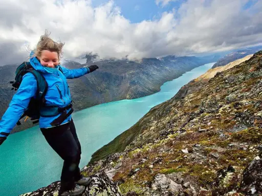 En kvinna med ryggsäck balanserar på en klippkant med en turkos sjö och berg i bakgrunden, en vy som påminner om Besseggen i Norge.