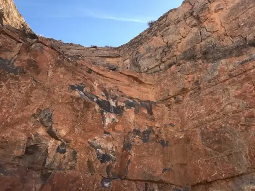 Acantilado de roca rojiza con vetas oscuras, mostrando diferentes tipos de canteras y la estratificación geológica bajo un cielo azul.
