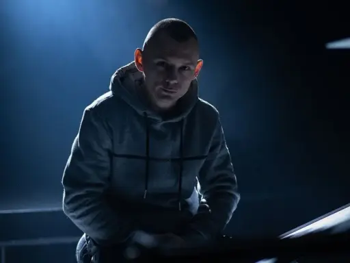 Młody mężczyzna w szarej bluzie z kapturem, z ogoloną głową, wygląda jak jeden z popularnych raperów, gotowy do występu.