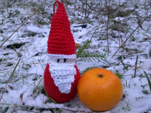 En virkad tomteluva med vitt skägg står i snön bredvid en apelsin.
