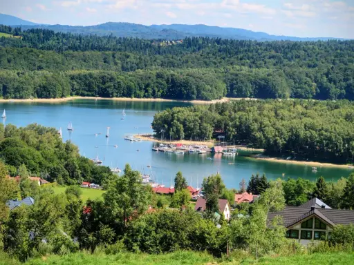 Malowniczy widok na jezioro z żaglówkami, otoczone lasami. W oddali widać góry, a na pierwszym planie domki letniskowe. Idealne miejsce na wypoczynek nad wodą, przypominające solina plaża.