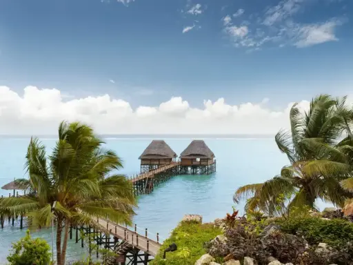 Dwa domki na wodzie połączone pomostem, otoczone palmami. Spokojne morze i błękitne niebo z chmurami. Zanzibar, pora deszczowa, ale pogoda dopisuje.
