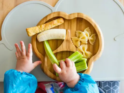 Babyhände greifen nach leckeren Baby-Snacks ab 6 Monaten: Banane, Gurke, Nudeln und ein Stück Brot auf einem Holztablett.