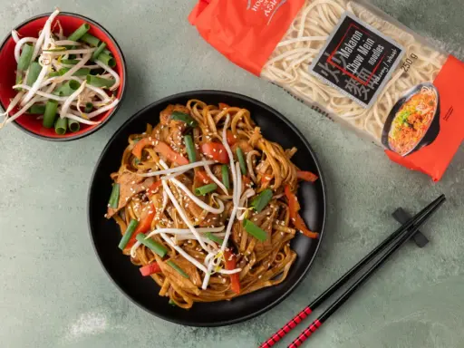 Pyszny makaron chow mein z warzywami i kiełkami fasoli na czarnym talerzu, obok paczka makaronu i miseczka kiełków.