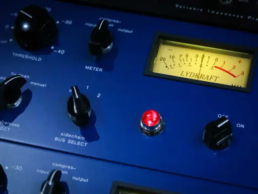 Gros plan sur un compresseur audio bleu avec des boutons, un VU-mètre et une lumière rouge allumée.