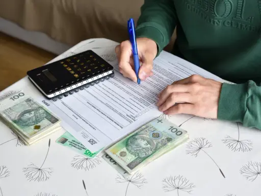 Osoba wypełnia dokumenty dotyczące umowy zlecenia i ubezpieczenia, obok leżą pieniądze i kalkulator.
