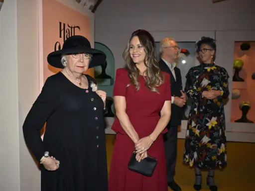 Prinsessan Sofia beundrar en utställning med hattar, där en äldre dam bär en elegant svart hatt.