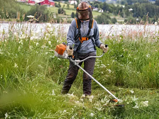 Persona con equipo de protección usa desbrozadora para cortar hierba alta, mostrando la diferencia entre desbrozadora y cortabordes en un paisaje rural.