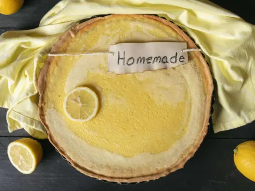 Ein hausgemachter Zitronenkuchen mit einem Zitronenschnitz und einem "Homemade"-Etikett, umgeben von gelben Tüchern und Zitronen.