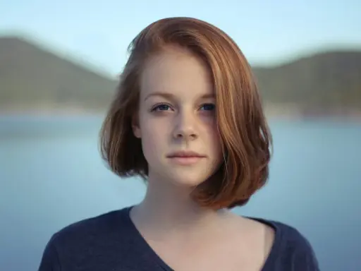 Ritratto fotografia di una giovane ragazza con capelli rossi e occhi azzurri, sullo sfondo di un lago e colline.