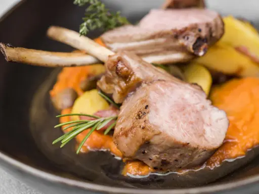 Chuleta de ternasco jugosa, servida sobre puré de zanahoria y patatas asadas, decorada con romero y tomillo.