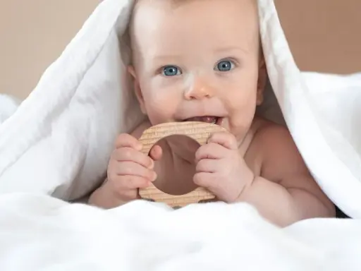 Ein Baby mit blauen Augen kaut auf einem Holzring. Vielleicht ist das der erster zahn baby, der durchbricht.