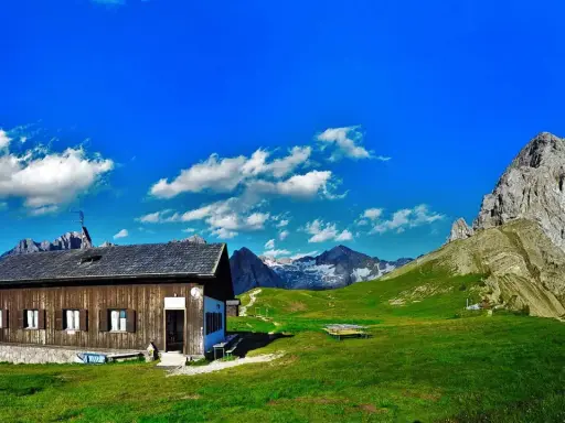 Rifugio Passo San Nicolò, un'accogliente baita in legno con vista su maestose cime innevate e un cielo azzurro brillante.