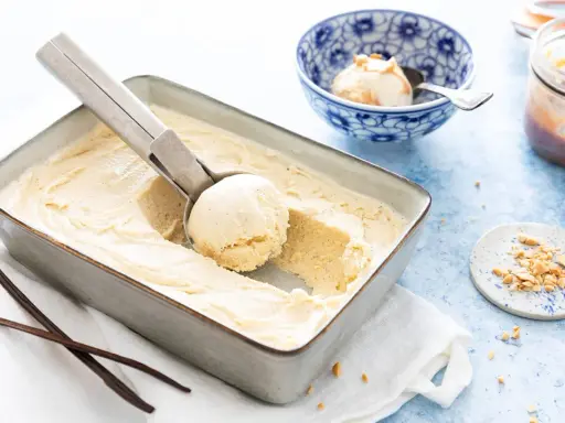 Une cuillère prélève une boule d'une glace vanille maison crémeuse dans un plat. Des gousses de vanille et des éclats de cacahuètes sont à proximité.