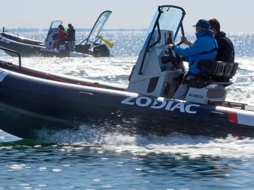 Deux bateaux Zodiac naviguent sur l'eau. L'un d'eux, au premier plan, porte le logo "ZODIAC" et semble communiquer sur le canal 9 VHF.