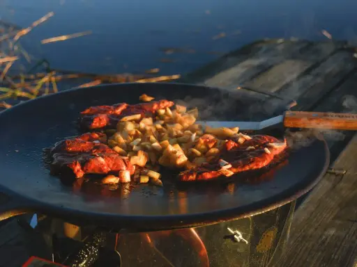 Kött och potatis steks på en rostig muurikka vid vattnet. En stekspade med trähandtag ligger bredvid.